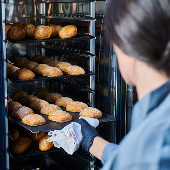 Eine Person entnimmt mit Handschuhen und Tuch frisch gebackene Brötchen aus dem CHEF'S COMBI von Hobart – mehrere Bleche mit goldbraunen Brötchen in einem professionellen Backofen.