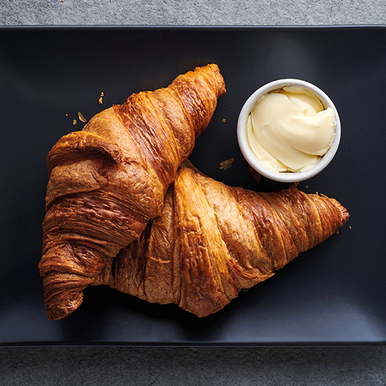 Zwei goldbraune, frisch gebackene Croissants auf schwarzem Teller mit einer Schale Butter – zubereitet mit dem CHEF'S COMBI von HOBART für perfekte Textur und Bräunung.