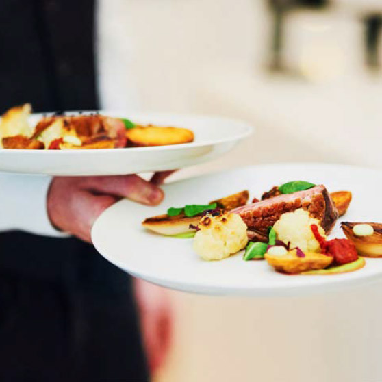 Eine Person hält zwei elegant angerichtete Teller mit Fleisch, geröstetem Gemüse und feinen Garnituren – zubereitet mit dem CHEF'S COMBI von HOBART für höchste Ansprüche in der Profiküche.