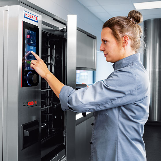 Eine Person bedient das Touchscreen-Display eines CHEF'S COMBI Geräts – sichtbar sind Zeit- und Temperatureinstellungen wie „30“ und „150°“, die eine präzise Steuerung im professionellen Küchenalltag ermöglichen.
