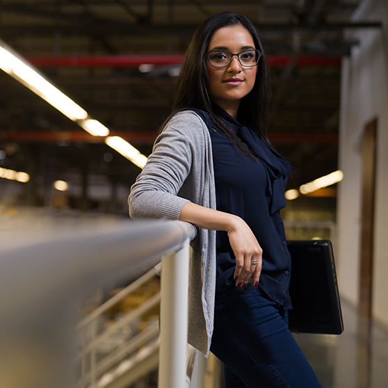 Person steht in einem modernen Industrie- oder Produktionsbereich und lehnt sich an ein Geländer, hält einen Laptop unter dem Arm. Im Hintergrund sind helle Beleuchtung und Strukturen der Halle sichtbar.