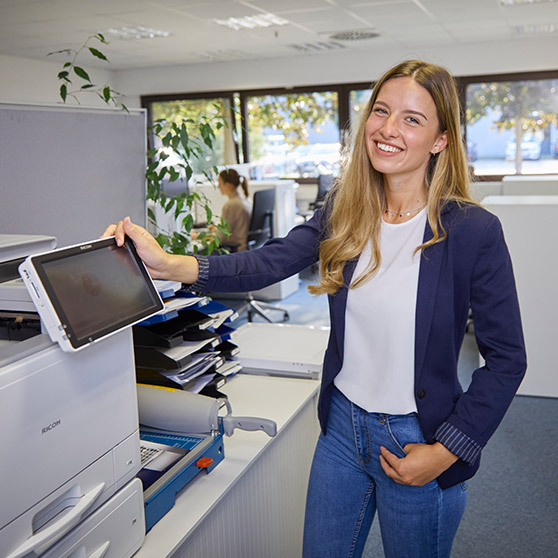 Frau im modernen Büro bedient einen Drucker zum Scannen und Drucken von Dokumenten. Im Hintergrund befinden sich helle Arbeitsplätze mit großen Fenstern, die viel Tageslicht hereinlassen.