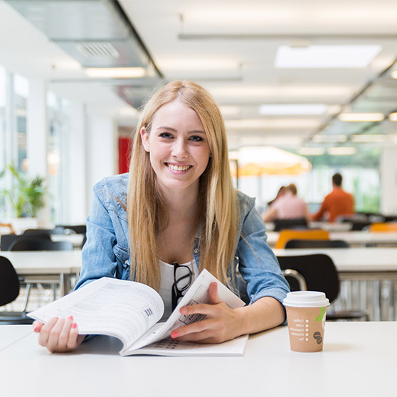 Person sitzt in einem hellen, modernen Raum und liest in einem aufgeschlagenen Buch. Auf dem Tisch liegt ein Kaffeebecher und eine Brille, im Hintergrund sind weitere Sitzplätze und große Fenster sichtbar.