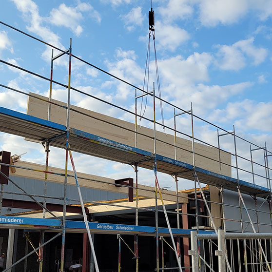 Baustelle mit Gerüst und Kran, der einen großen Holzträger auf die Stahlkonstruktion hebt. Im Hintergrund blauer Himmel mit Wolken.