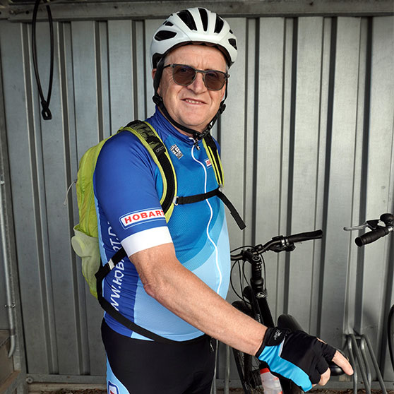 Person in blauer Fahrradbekleidung mit Helm und gelbem Rucksack steht neben einem schwarzen Fahrrad in einer Fahrradgarage. Auf dem Trikot ist das HOBART-Logo sichtbar.