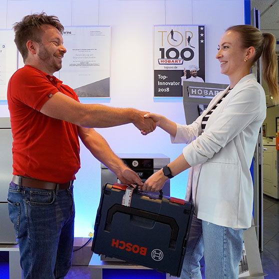 Zwei Personen in einem modernen Arbeitsumfeld geben sich die Hand bei einer Übergabe. Im Vordergrund ein schwarzer Werkzeugkoffer, im Hintergrund Poster und Auszeichnungen, darunter „Top 100 – Top Innovator 2018“.