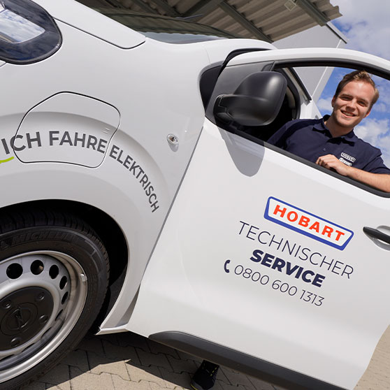 Elektro-Servicefahrzeug mit der Aufschrift „ICH FAHRE ELEKTRISCH“ und „HOBART Technischer Service“ sowie Telefonnummer 0800 600 1313, geparkt unter einem Dach bei sonnigem Wetter.