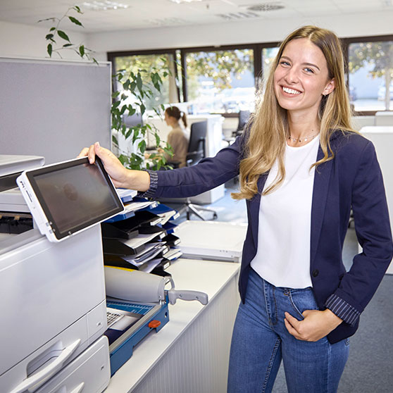 Helles, modernes Büro mit einer Person, die einen Drucker bedient. Auf dem Schreibtisch liegen Papierstapel und Büromaterial, im Hintergrund große Fenster mit Tageslicht und weitere Arbeitsplätze.