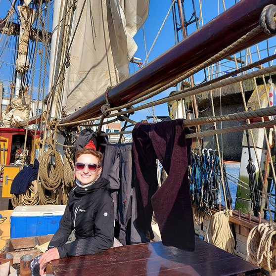 Traditionelles Segelschiff im Hafen mit eingerollten Segeln, Seilen und Kleidung zum Trocknen. Eine Person sitzt am Holztisch unter blauem Himmel.