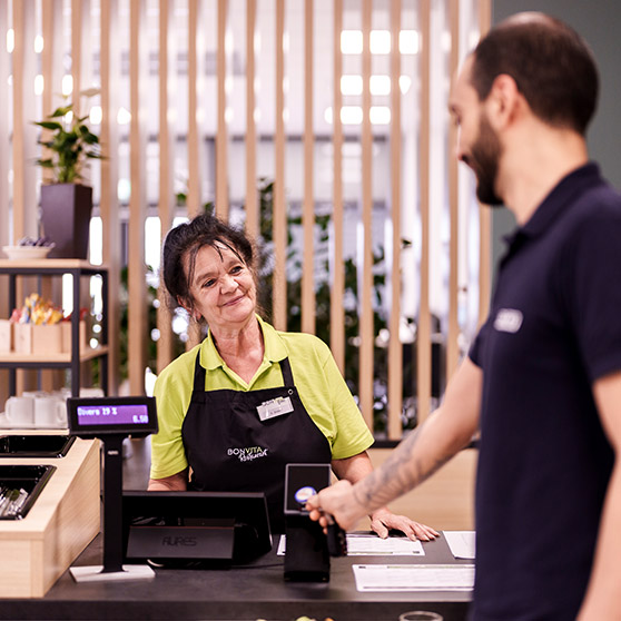 Bezahlvorgang in einer modernen Betriebsgastronomie: Ein Gast nutzt kontaktlose Kartenzahlung am elektronischen Kassenterminal. Der Betrag wird auf einem digitalen Display angezeigt. Im Hintergrund sorgen Holzlamellen und grüne Pflanzen für ein einladendes Ambiente.