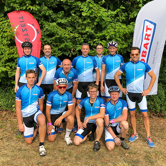 Gruppe von Radfahrern in HOBART-Trikots bei einem Teamevent – Aufnahme vor grüner Hecke mit HOBART-Banner und Sportausrüstung.