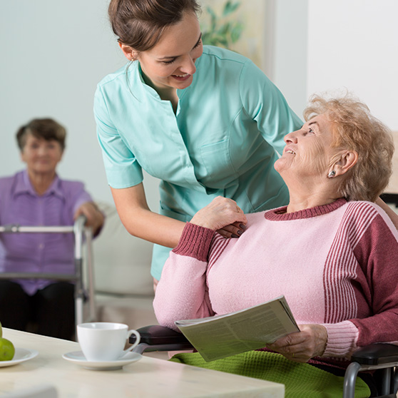 Pflegekraft unterstützt eine ältere Person im Rollstuhl beim Lesen einer Zeitung in einem hellen Pflegeheim, mit einer Tasse Kaffee und Obst auf dem Tisch, weitere Bewohner im Hintergrund.