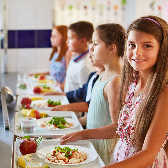 Reihe von Kindern in einer Schulkantine mit Tabletts, darauf Teller mit Nudeln, Salat, frischem Obst wie Äpfeln und Bananen sowie Gläsern Wasser, im Hintergrund helle Wände und eine Essensausgabe.