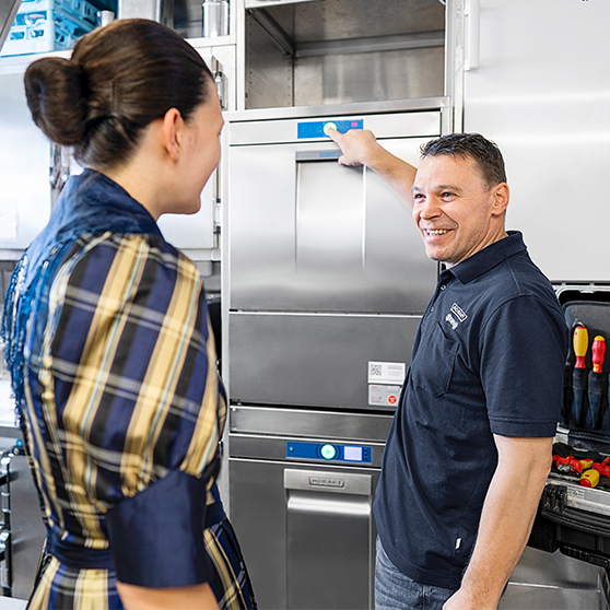 HOBART Servicetechniker demonstriert die Bedienung einer professionellen HOBART Spülmaschine in einer Gastronomieküche, mit Werkzeugkoffer und Edelstahltechnik im Hintergrund.