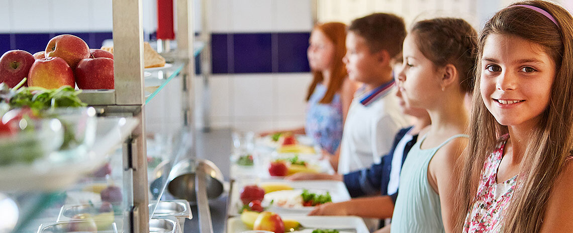 Essensausgabe im Schulcatering mit mehreren Personen, die an einer Theke mit Obst und vorbereiteten Mahlzeiten anstehen.
