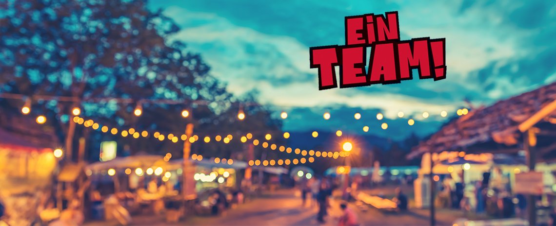 Eine Außenaufnahme eines Veranstaltungsbereichs mit dekorativen Lichterketten, mehreren Ständen im Hintergrund und dem auffällig platzierten Grafiktext EIN TEAM! im oberen Bildbereich.