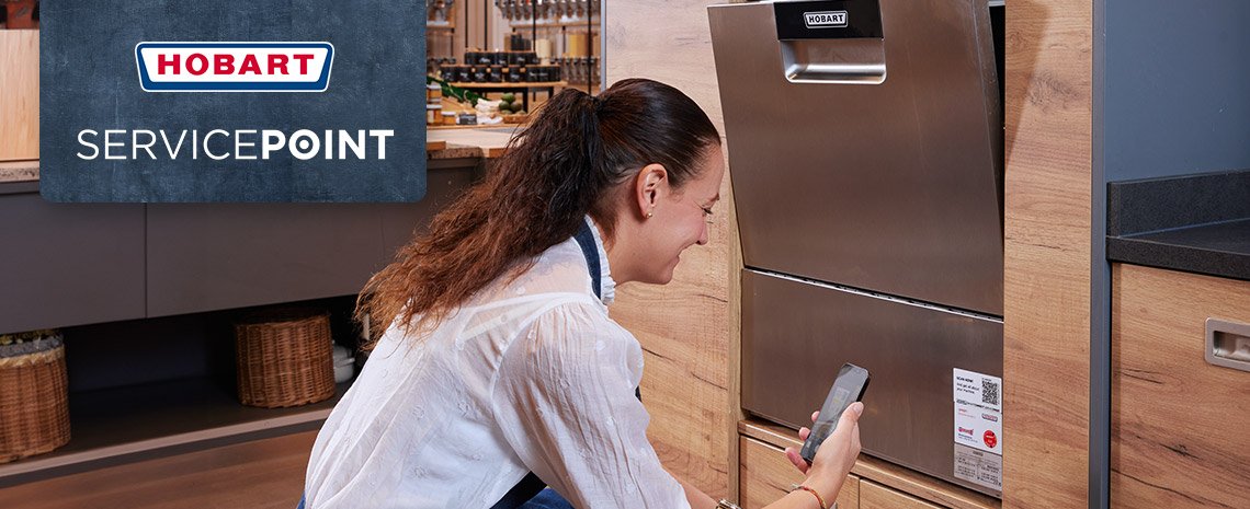 Eine Person kniet vor einer eingebauten HOBART Spülmaschine und hält ein Smartphone, während im Hintergrund ein SERVICEPOINT Schild zu sehen ist.