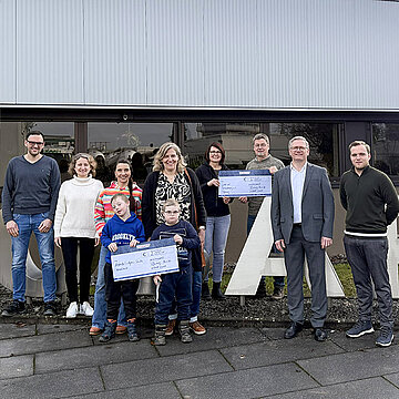 Eine Gruppe von Personen steht vor einem Gebäude und hält große Spendenschecks, begleitet von mehreren Kindern, stellvertretend für die Übergabe von Mitteln aus einer von HOBART Mitarbeitenden unterstützten Spendeninitiative an regionale Einrichtungen.