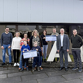 Eine Gruppe von Personen steht vor einem Gebäude und hält große Spendenschecks, begleitet von mehreren Kindern, stellvertretend für die Übergabe von Mitteln aus einer von HOBART Mitarbeitenden unterstützten Spendeninitiative an regionale Einrichtungen.